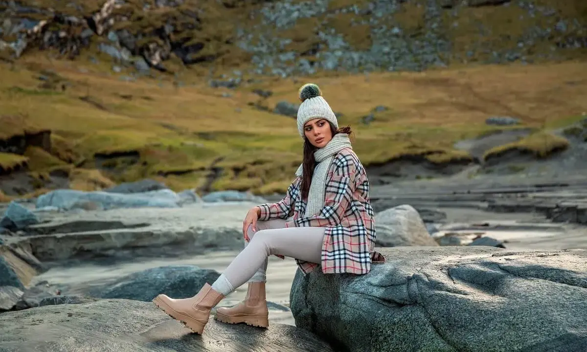 Modna kobieta w czapce i szaliku prezentuje stylizacje z kurtką koszulową w kratę, siedząc na kamieniach nad wodą.