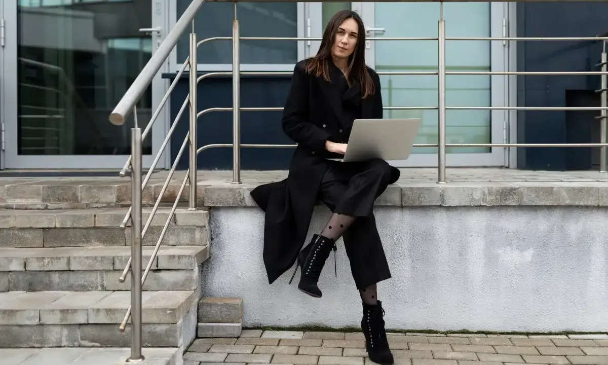 Elegancka kobieta w czarnym płaszczu i spodniach, z laptopem na kolanach, siedzi na schodach. Stylowe czarne płaszcze to podstawa wielu udanych stylizacji.