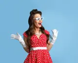 Kobieta w czerwonej sukience w grochy, białych rękawiczkach i okularach w stylu pin up, z uniesionymi rękami, wyraża radość.