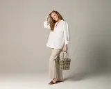 Eleganckie spodnie lniane damskie stylizacje: luźna biała koszula, beżowe spodnie, pleciona torebka i brązowe klapki.