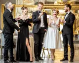 Elegancka czwórka w strojach black tie wznosi toasty. Kobieta w czarnej sukni z rozcięciem i mężczyźni w smokingach.