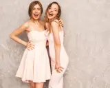 Dwie uśmiechnięte kobiety w pastelowych sukienkach, idealne na różne rodzaje dress code'u, od koktajlowego po wieczorowy.