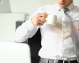 Mężczyzna trzyma kubek z kawą, a na jego białej bluzce widoczna jest duża plama z kawy.