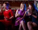 Trzy kobiety w barze podczas przyjęcia koktajlowego. Jedna rozmawia przez telefon, dwie pozostałe plotkują przy drinkach.