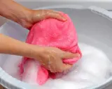 Delikatne pranie wełny merino w pianie. Ręce ugniatają różowy materiał w misce z wodą.