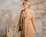 Elegancka kobieta w płaszczu w kolorze camel, sukience w kwiaty i okularach przeciwsłonecznych pozuje przy starej walizce.