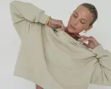 Kobieta w bluzie w kolorze oliwkowym, z złotą biżuterią, delikatnie poprawia dekolt.