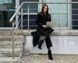 Elegancka kobieta w czarnym płaszczu i spodniach, z laptopem na kolanach, siedzi na schodach. Stylowe czarne płaszcze to podstawa wielu udanych stylizacji.