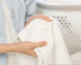 Dłonie trzymają białą tkaninę z plamami, pokazując problem, jak usunąć podkład z kołnierza kurtki.