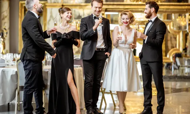 Elegancka czwórka w strojach black tie wznosi toasty. Kobieta w czarnej sukni z rozcięciem i mężczyźni w smokingach.