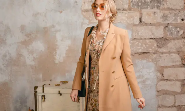 Elegancka kobieta w płaszczu w kolorze camel, sukience w kwiaty i okularach przeciwsłonecznych pozuje przy starej walizce.