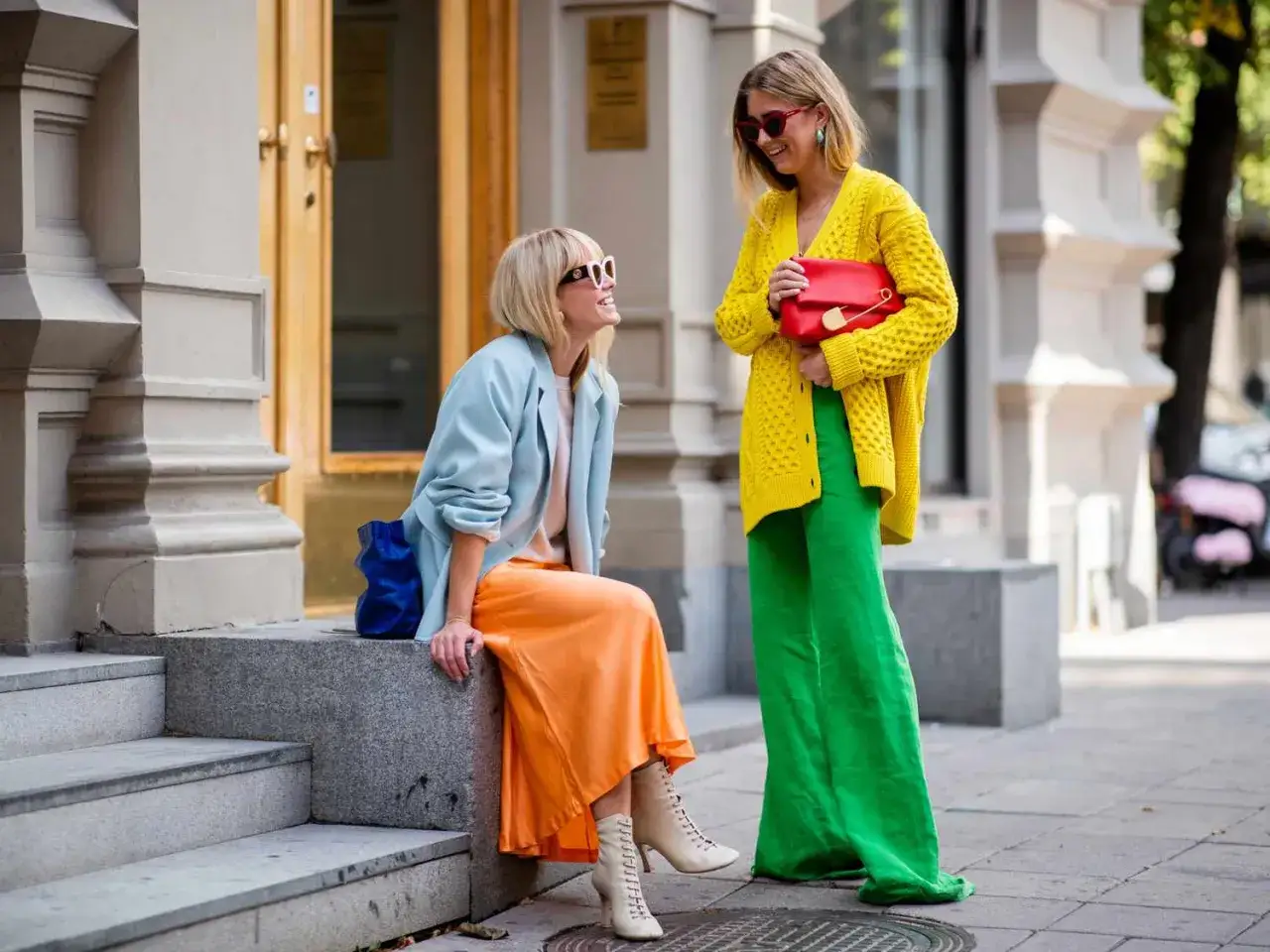 Dwie kobiety prezentują odważny styl color blocking: jedna w błękitnej marynarce i pomarańczowej sp&oacute;dnicy, druga w ż&oacute;łtym kardiganie i zielonych spodniach.