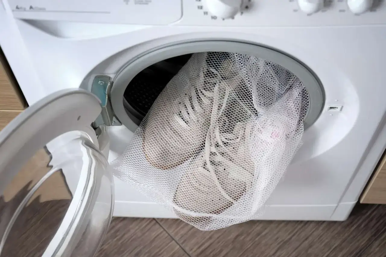 Białe buty sportowe w siatkowej torbie czekają na pranie w pralce. To prosty spos&oacute;b, jak prać buty, by odzyskały świeżość.