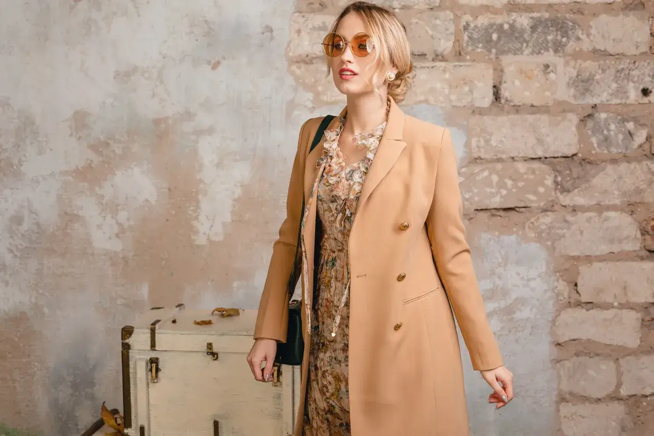 Elegancka kobieta w płaszczu w kolorze camel, w sukience w kwiaty, z okularami przeciwsłonecznymi.