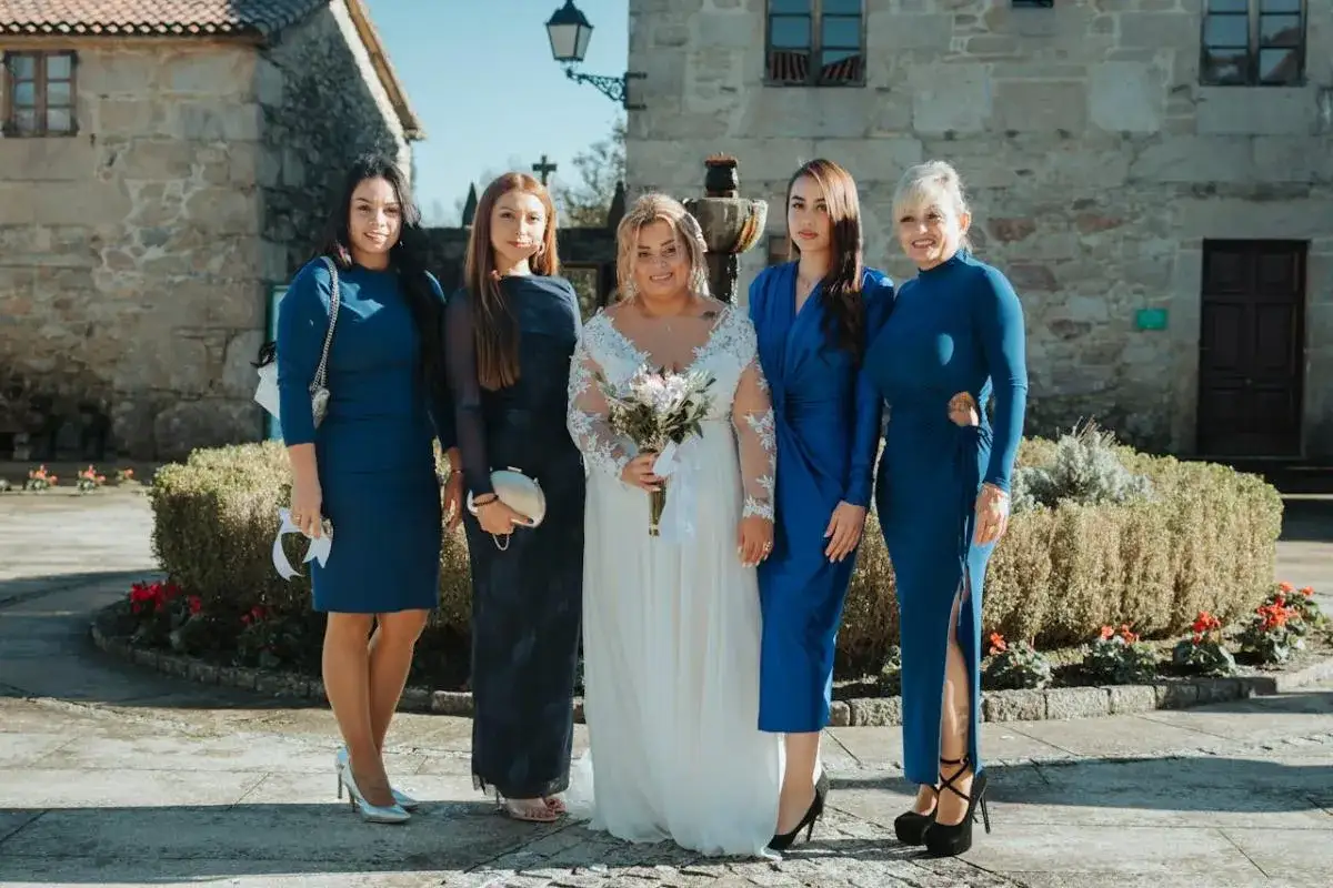 Panna młoda w białej sukni i jej druchny w eleganckich kreacjach w kolorze chabrowym pozują do zdjęcia na tle kamiennego budynku.