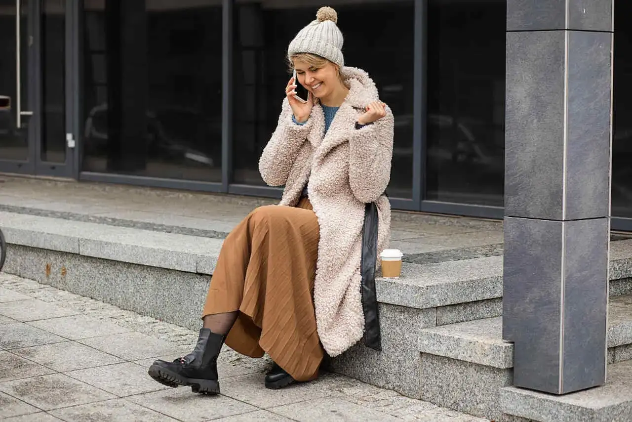 Uśmiechnięta kobieta w beżowym płaszczu z futerkiem i brązowej sp&oacute;dnicy rozmawia przez telefon, siedząc na schodach.
